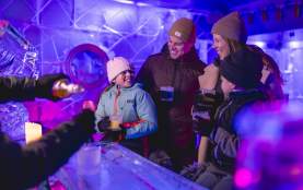 A family of four enjoying drinks in an ice bar, surrounded by illuminated ice sculptures and glowing purple and blue lighting. They are dressed warmly in coats, hats, and gloves, smiling and sharing a joyful moment.