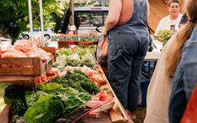 Visitors browse a sunny outdoor farmers’ market stall filled with fresh local produce, including leafy greens, carrots, and squash, under white canopy tents.