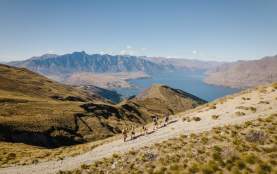 A group of hikers climbing the top of a mountain with views of Lake Whakatipu and Remarkable Mountains in the background