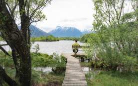 Man sits on a seat overlooking a lake, surrounded by green trees with a snowcapped mountain in the background
