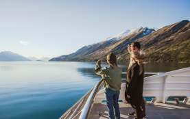 Three people standing on the deck of a boat, gazing out at the calm blue waters of a lake surrounded by mountains. The landscape features snow-capped peaks and lush greenery under a clear blue sky, creating a serene and picturesque setting