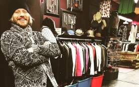 A smiling man wearing a black beanie and patterned grey sweater stands with his arms crossed inside a vintage clothing store, surrounded by racks of colourful jackets, hats, and accessories.