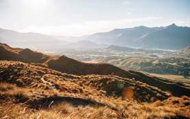 Far shot of three bikers descending a golden tussock-covered mountain with valley and mountain in the background