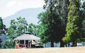 A small red pavilion sits among tall trees with mountains in the background on a sunny day.