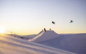Snowboarders and a skier perform aerial tricks off a jump at Cardrona Alpine Resort at sunrise.