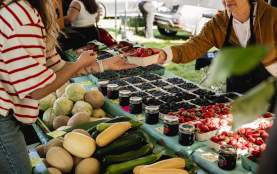 A vendor hands a basket of strawberries to a customer at a farmers’ market, with fresh produce and jars of preserves on display.