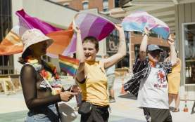 Three people walking on the street, each holding a different flag over their heads. From left to right- Pride flag, lesbian flag, and trans flag.