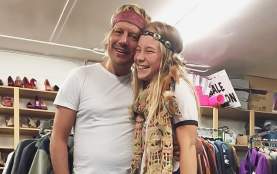 Two smiling people wearing headbands pose together inside a secondhand clothing store, surrounded by racks of jackets and shelves of shoes.
