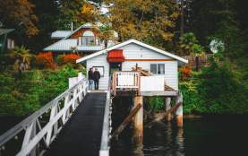 Two people stand on a narrow walkway leading to a small oceanfront house on stilts above the water, surrounded by trees and fall foliage.