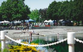 People gather along a waterfront park in Nanaimo (Maffeo Sutton Park), with children wading in the water and market tents and the ‘Nanaimo’ sign in the background.