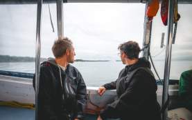 Two individuals sit inside a small boat, looking out over calm coastal waters toward a distant shoreline under an overcast sky.