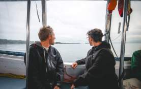Two individuals sit inside a small boat, looking out over calm coastal waters toward a distant shoreline under an overcast sky.