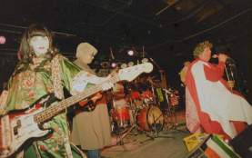 Photo of the band Swine King performing on stage a woman is standing on the left wearing a green floral dress and playing bass guitar a man is standing behind her wearing a long brown jacket and fuzzy tan hat and playing the electric guitar and lead singer Randy Biscuit Turner is standing on the right wearing a red toga outfit and singing into a microphone