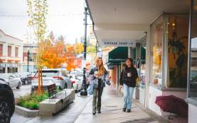Two people stroll past shops on a rainy autumn day in downtown Nanaimo, holding coffee as colourful fall trees line the street.