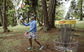 A person plays disc golf in a wooded park, tossing a disc toward a metal basket surrounded by trees.