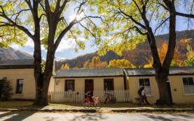 Historic miners’ cottages in Arrowtown dappled in autumn sunlight, framed by golden trees, with red bikes parked out front and a couple walking a dog beneath the canopy.