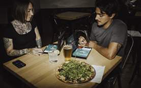 Two people play a tabletop game at a restaurant table with pizza and drinks in front of them.