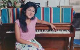 Historic photo of Tish Hinojosa sitting on a piano bench and facing the camera with her left arm resting on the piano