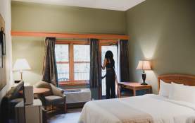 A cozy hotel room with a bed, desk, and seating area as a person opens a balcony door to let in natural light.