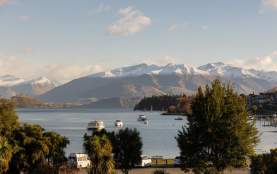 Peaceful view over Lake Wānaka with boats anchored on the still water, framed by greenery and snow-dusted mountains under a clear afternoon sky.
