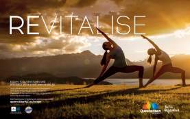 Two people are practising yoga outdoors at sunset on a grassy hill, with mountains and a lake in the background. The text reads “REVITALISE” and promotes a pre-winter escape to Queenstown, New Zealand.