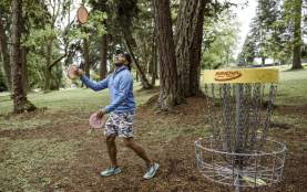 A person plays disc golf in a wooded park, tossing a disc toward a metal basket surrounded by trees.