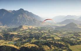 Person paragliding over a green valley with the Remarkables mountain range in the background