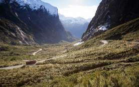 Wide shot of a bus traveling on the road to Milford Sound surrounded by towering snow-capped mountains and native bush