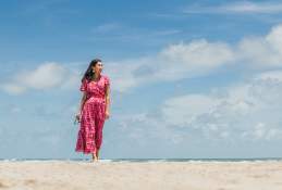 A woman in a pink and red dress walks along the beach