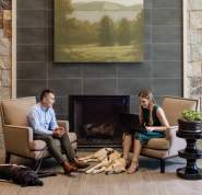 Two people are seated in front of a fireplace with their service dogs lying asleep at their feet. On person has a laptop open and is describing her report to the listener.