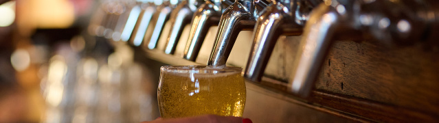 Close-up of a bartender pouring a golden beer from a row of draft taps labeled with Milwaukee Brewing Company beers into a small tasting glass.
