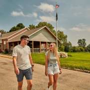 Two people, in summer, walking down a street with a VFW building behind them, and rolling hills.