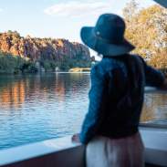A woman enjoying the scenery on an Ord River cruise - the woman is standing on the vessel looking across the water at a cliff faced bathed in sunlight.