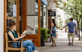 A woman sits on a bench reading a book while a couple in the background is walking into a shop.
