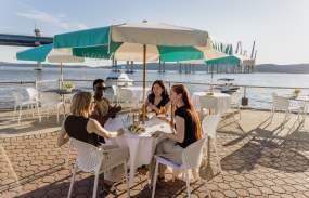 A group of four people sit at an outdoor dining table overlooking the Hudson River. They are laughing and chatting.