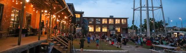 A crowd gathers outside Lowe Mill ARTS & Entertainment in Huntsville during an evening outdoor concert, with string lights glowing along the historic brick building and the iconic water tower rising above the lawn as families and visitors enjoy live music at sunset.
