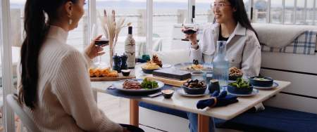 Two women clinking wine glasses in a restaurant overlooking the sea.