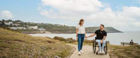 A man in a wheelchair holding hands with a standing woman on a section of the coast path with the sea in the background.