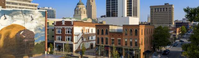 Aerial view of The Landing in downtown Fort Wayne at sunset, showcasing historic buildings, tree-lined streets, and the city skyline in the background.