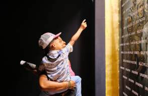 African American man holding a young child in front of the plaque with engraved names of the Clotilda, the Last American Slave Ship