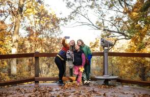 Family smiles and poses for a selfie outdoors during a fall day.
