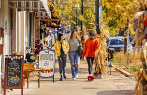 Three women shopping in Depot Town, walking along storefronts on a sidewalk during the fall season. Yellow and orange leaves in the background.