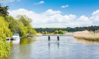 Two paddle boarders on the River Arun, with a boat moored to the left and grassy bank to the right, in Sussex