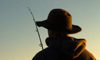 Photograph showing a person fishing from behind, with the dusk light only just illuminating them.