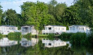 White, modern lodges are dotted along the shore of a lake and surrounded by trees at Hoburne Cotswold in the Cotswolds