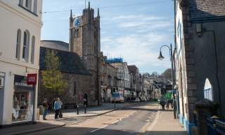 A quaint street scene features a stone church with a tall clock tower, flanked by shops and buildings; people walk under a bright blue sky.