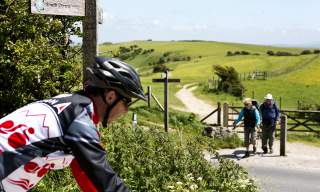 A mountain biker rides along the South Downs Way, with walkers on a trail in the background