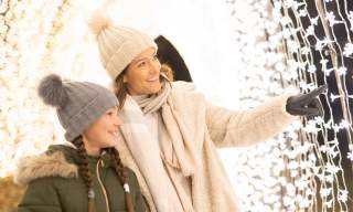 A mother and daughter wearing wooly hats, scarves and gloves in a tunnel of fairy lights