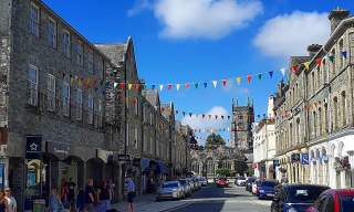 A vibrant street scene with colorful pennant flags strung between gray stone buildings under a bright blue sky. People and parked cars line the street.