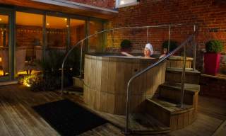 A lady relaxes at night in an outdoor wooden hot tub, surrounded by plants and subdued lighting at The Greenway Hotel & Spa in the Cotswolds